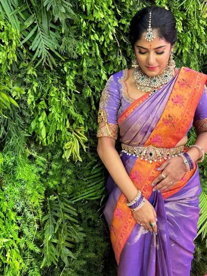 A client wearing a purple Kanchipuram saree with a vibrant orange border. Her custom purple blouse is heavily embroidered, creating a rich and colorful ensemble.