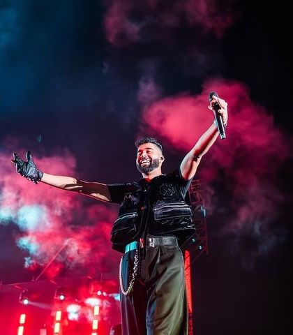 AP Dhillon owning the stage, arms outstretched, with plumes of colored smoke creating a dramatic backdrop.