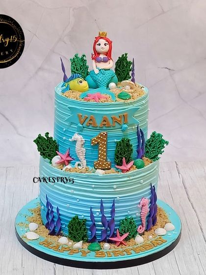 This two-tier mermaid cake for a first birthday features a beautiful blue frosting, a fondant mermaid topper, and edible decorations like seahorses, starfish, and seaweed.