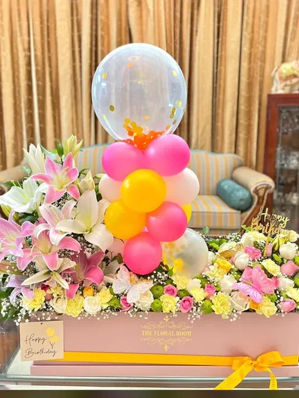 A large and luxurious birthday flower box. This arrangement is filled with pink lilies, yellow roses, and other pastel flowers, and is topped with a fun balloon cluster.