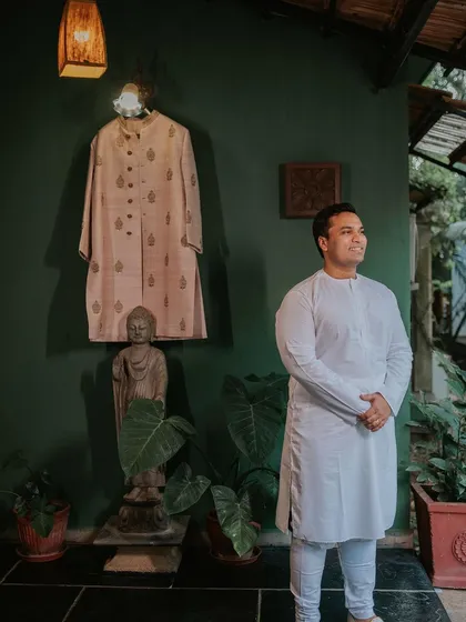The groom, getting ready, stands next to his wedding sherwani, which is hung艺术fully against a green wall.
