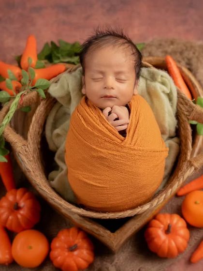 An autumn harvest theme featuring a newborn wrapped in orange, nestled in a basket surrounded by carrots and pumpkins.