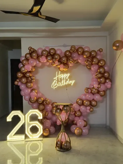 A stylish 26th birthday setup featuring a heart-shaped balloon arch in rose gold and pink. The look is completed with a "Happy Birthday" neon sign and large marquee numbers.