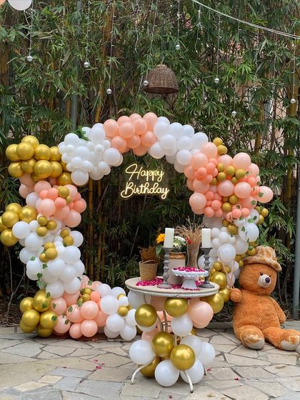 A beautiful boho-chic inspired balloon ring for an outdoor birthday party. The arch of peach, white, and gold balloons is set against a natural bamboo background, with a small cake table and a teddy bear prop for a whimsical touch.