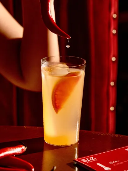 A spicy chili cocktail against a dramatic red background. The single drop of chili essence adds a touch of action and hints at the heat within.