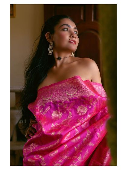 A soft, romantic portrait using natural window light. The model, draped in a pink silk saree, is captured in a moment of thought, highlighting the fabric's texture and the mood of the scene.