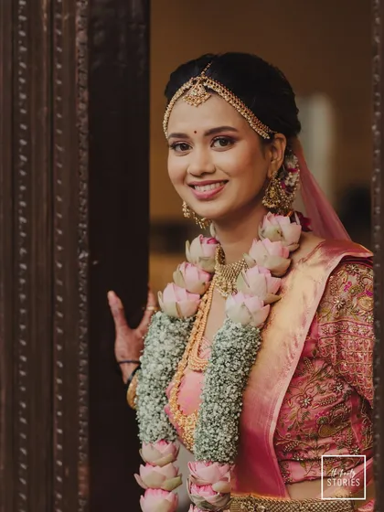 A happy bride peeking through a doorway, her face framed by a beautiful lotus garland. Her bright smile captures the excitement of her wedding day.