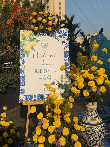 A charming welcome easel for the Haldi, featuring a lemon and tile motif, surrounded by bright yellow marigolds in ornate blue and white vases.