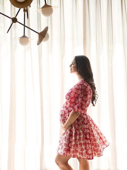 Bathed in the soft light of a window, the mother-to-be stands in profile, her red and white dress adding a pop of color to this serene, at-home portrait.