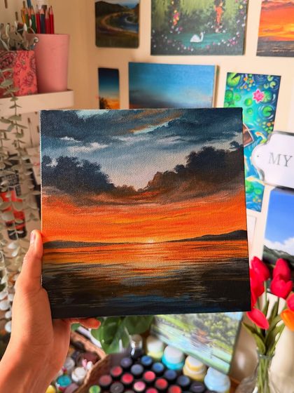Holding up my finished orange sunset painting in my studio, with my other artworks visible in the background.