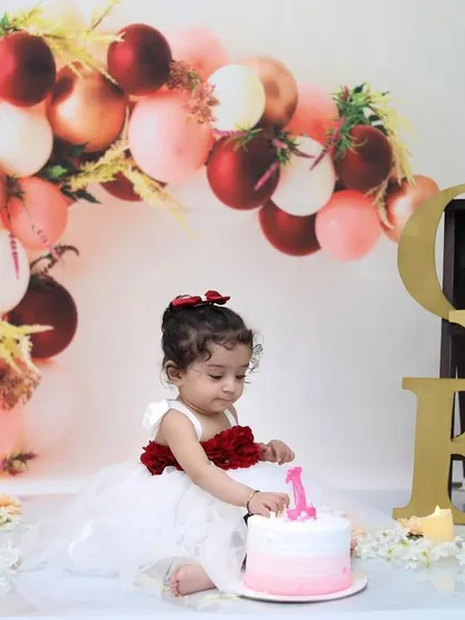 A little girl in a beautiful white and red dress delicately touches the "1" candle on her pink ombre cake during her elegant first birthday photoshoot.