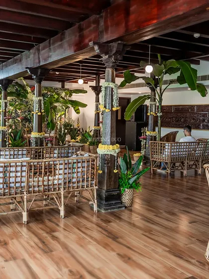 A traditional South Indian Haldi setup. The space features dark wood pillars, cane furniture, and is filled with banana plants and marigold garlands, creating a warm and authentic atmosphere.