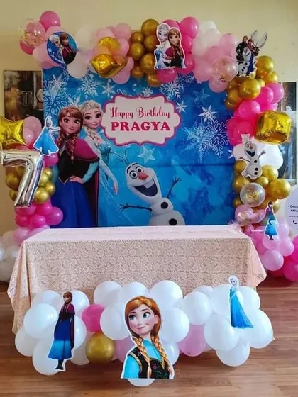 A 'Frozen' themed birthday party with a backdrop featuring Elsa, Anna, and Olaf. The setup includes character cutouts and a balloon garland.