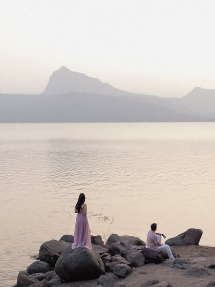 A wide, atmospheric shot of a couple enjoying the serene landscape of Pawna Lake. The composition creates a sense of scale and tranquility, highlighting the beauty of nature as a backdrop for their love.