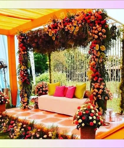 A colorful mandap-style seating for a Mehendi or Haldi function. The structure is draped in orange fabric and heavily decorated with multi-colored flowers for a celebratory feel.