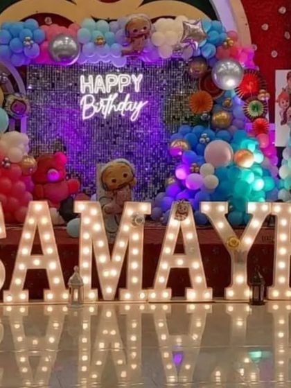 A massive stage setup for a birthday, featuring large light-up letters spelling "SAMAYRA" and a colorful, complex balloon backdrop with a neon sign.