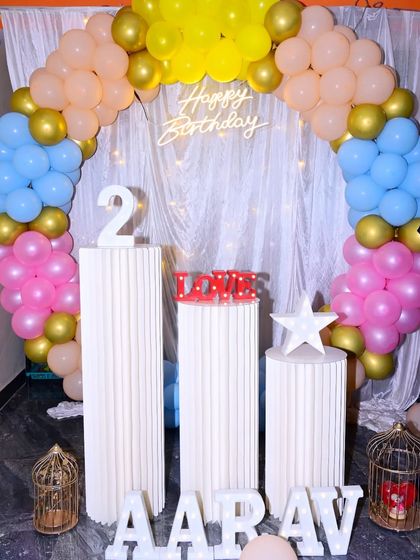 A second birthday celebration with a pastel blue, pink, and yellow balloon arch. The setup is completed with a 'Happy Birthday' neon sign and marquee letters.