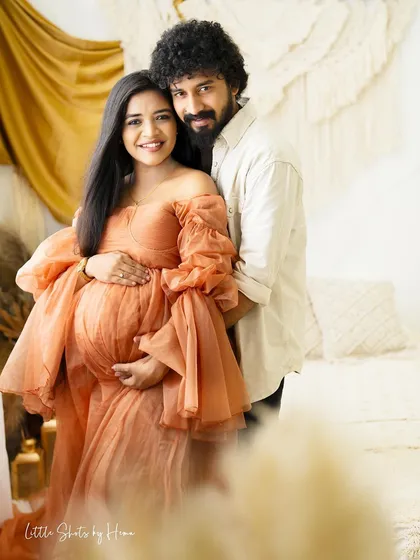 A smiling couple captured in a bright, boho-themed room. The partner stands behind the mom-to-be, who is wearing a lovely peach gown, both looking happily towards the camera.