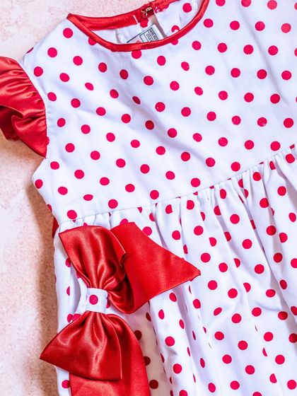 This playful dress features red polka dots on a white background. The ruffled cap sleeves and a large, dramatic bow on the side make it a fun and festive choice.