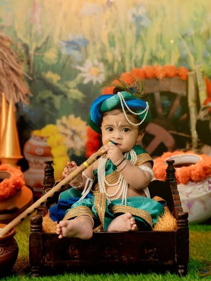 A baby boy dressed as Little Krishna, playfully tasting "butter" from a pot in a rustic village setting.
