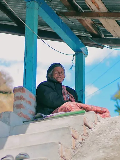 An elderly woman sits peacefully on the steps of her porch in a Himachali village. The simple, rustic architecture and the clear blue sky in the background frame a portrait of contentment and a life connected to the mountains.