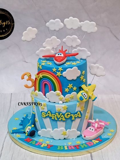 A high-flying two-tier airplane-themed cake for a 3rd birthday. The sky-blue cake is decorated with fondant clouds, a rainbow, and characters from the "Super Wings" show.