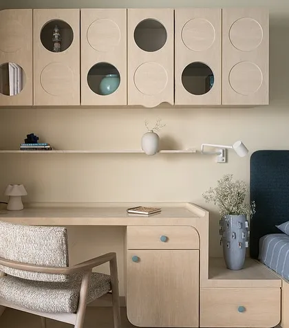 This son's bedroom in the 'Soft Serve' project uses soft blue tones and light wood to create a calm and playful atmosphere. The custom desk and overhead storage unit feature circular cutouts, adding a touch of modern geometry.