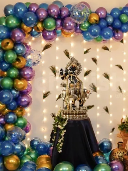 A stunning Janmashtami balloon arch in metallic shades of blue, purple, and green, with peacock feathers accenting the wall behind the Krishna idol.