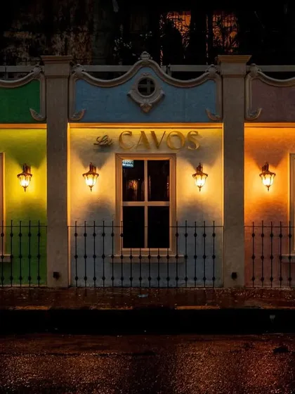 The exterior facade of Los Cavos at night, designed to look like a row of colourful colonial buildings. Each section is painted a different hue and lit by traditional wall sconces, creating a vibrant and welcoming entrance that sets the tone for the experience inside.