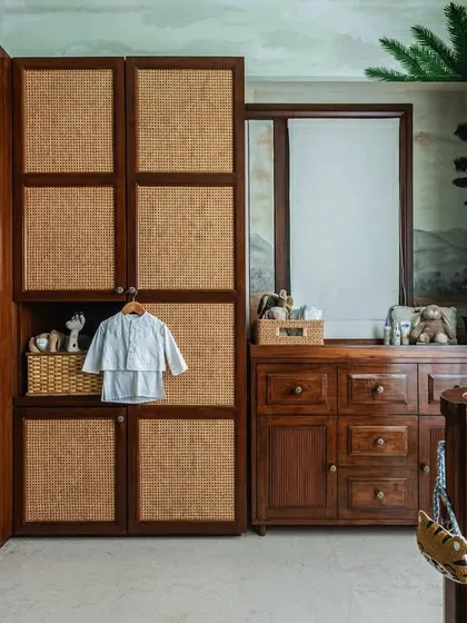 Functionality is key in a nursery. Here you can see the combination of rattan and wood cabinetry, providing ample storage, alongside a classic wooden changing table and a retro-style Smeg fridge for convenience.