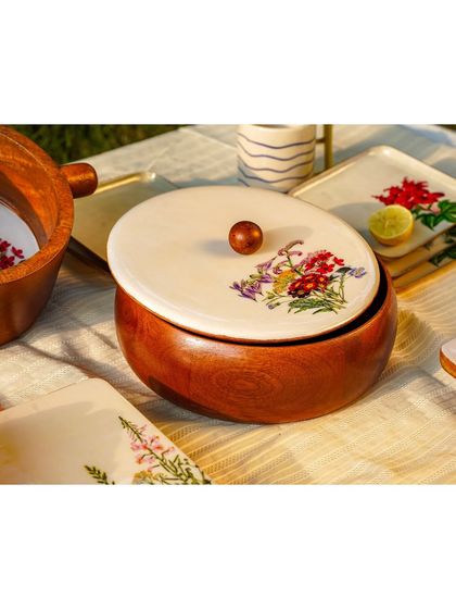 A detailed shot of a wooden serving bowl with a painted lid, part of an outdoor picnic scene. The focus is on the unique craftsmanship of the product.