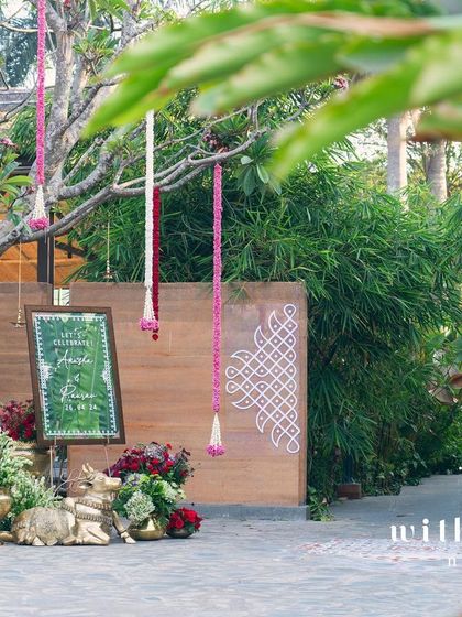 The entrance to a summer wedding, decorated with hanging floral garlands and a welcome sign featuring a traditional kolam design.