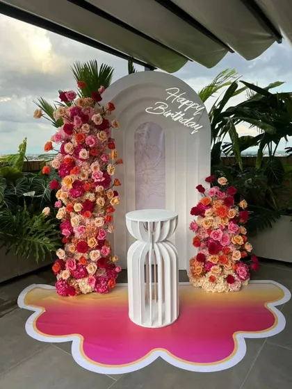 An elegant and modern birthday backdrop combining fresh flowers and custom structures. This decor features a white arched panel with a neon "Happy Birthday" sign, flanked by stunning pink and red floral towers, perfect for a sophisticated celebration.