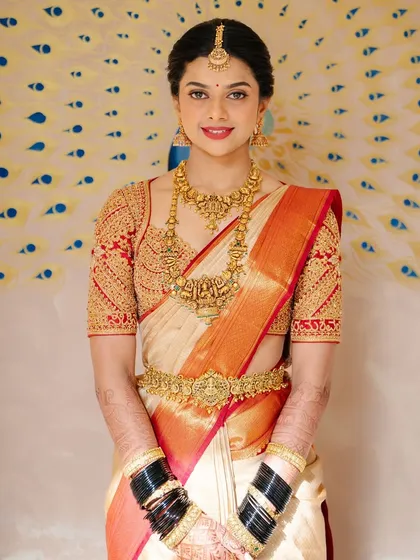 Bride Supritha radiates in a mesmerizing off-white Kanchivaram saree, complemented by a red blouse hand-crafted with zari, zardosi, and sequins.