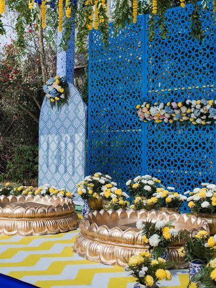 A close-up of the blue and yellow Haldi decor, showing the traditional brass urlis filled with yellow and white flowers against a jaali-patterned backdrop.