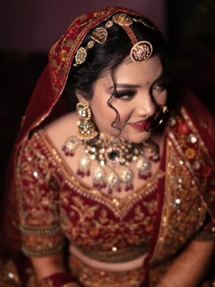 A close-up portrait of a bride, her expression soft and thoughtful. The dark background emphasizes the rich colors of her lehenga and jewelry.