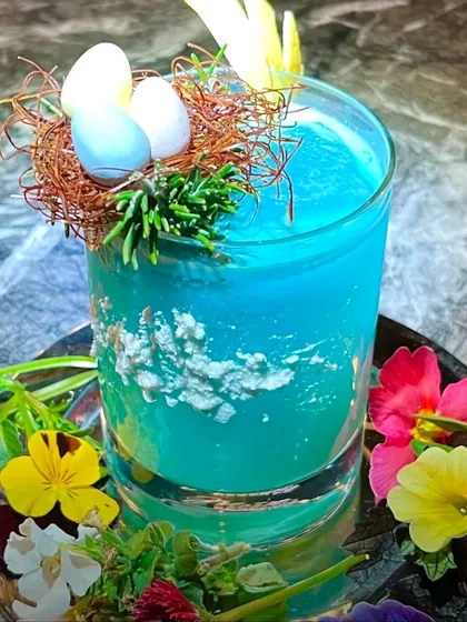 An Easter-themed blue mocktail. The creative garnish, featuring a small nest with candy eggs, shows how we can tailor our drinks to any event theme.