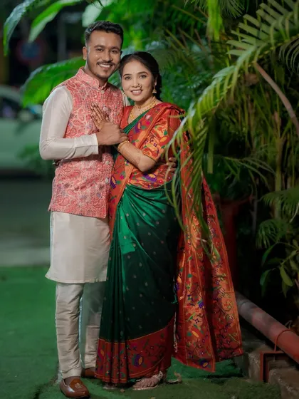 A full-length portrait of the couple, showcasing the bride's stunning green and red Paithani saree against a backdrop of lush palm leaves.