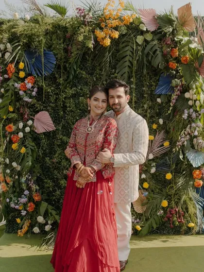 A happy couple posing in front of a lush green photo wall, adorned with a variety of colorful and tropical flowers. It's a fresh and vibrant backdrop for a daytime event.