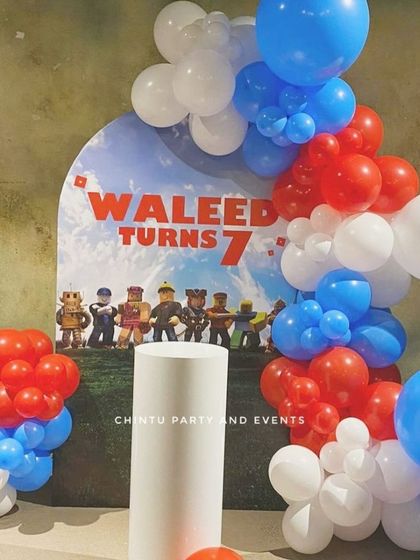 A Roblox themed 7th birthday decoration. The setup features a balloon arch in red, white, and blue around a personalized backdrop with game characters.