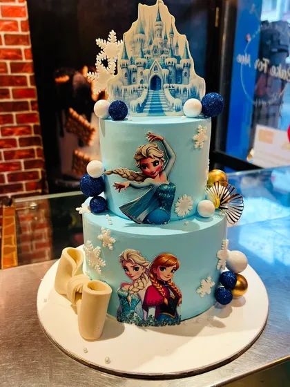 A two-tier Frozen-themed cake featuring Elsa, Anna, and Olaf, with a beautiful ice castle topper and snowflake decorations.