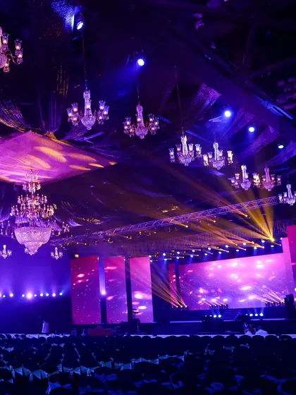 The calm before the storm of celebration. This wide shot shows the scale of a concert-style reception, with dozens of chandeliers suspended over the hall and a massive stage, all lit in dramatic purple and gold hues.
