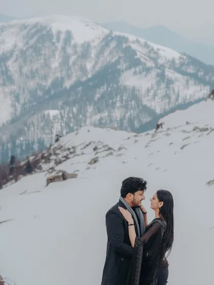 A couple stands close together, surrounded by snow-covered terrain and a backdrop of majestic white peaks, creating a stunning winter wonderland portrait.