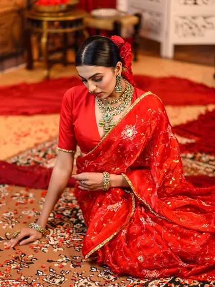 A beautiful composition of a model seated on a traditional rug. The rich red of the saree and the intricate patterns of the rug create a visually dense and luxurious image.