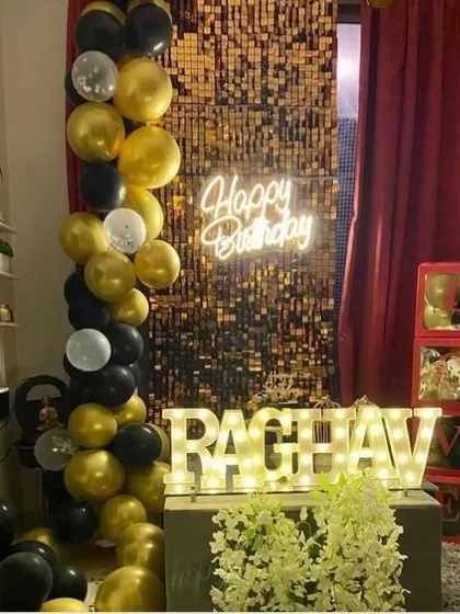 A sophisticated birthday setup for Raghav. A gold sequin wall is paired with a black and gold balloon garland, a neon sign, and illuminated marquee letters.