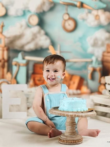 A happy little explorer enjoying his cake during an aviator themed first birthday shoot. The backdrop is filled with clouds, vintage planes, and clocks to create a sense of adventure.