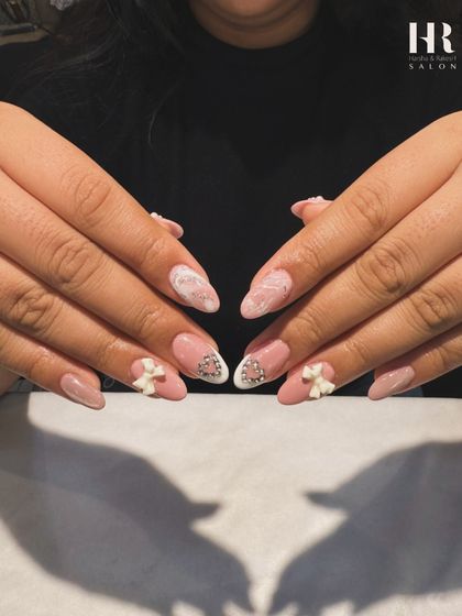 Sweet and delicate nail art. This design features a soft pink base with white marble accents, 3D bows, and sparkling heart-shaped gems, creating a romantic and feminine look.