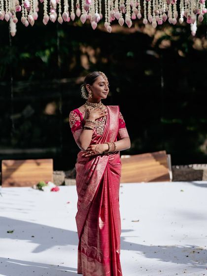 A bride stands on a white platform, her pink saree and the hanging floral decor creating a dreamy, ethereal portrait.