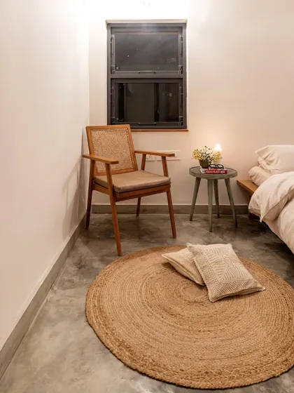 A cozy bedroom corner with a jute rug, a simple wooden chair, and soft, neutral bedding. The space feels calm and inviting, with a focus on natural materials and textures.