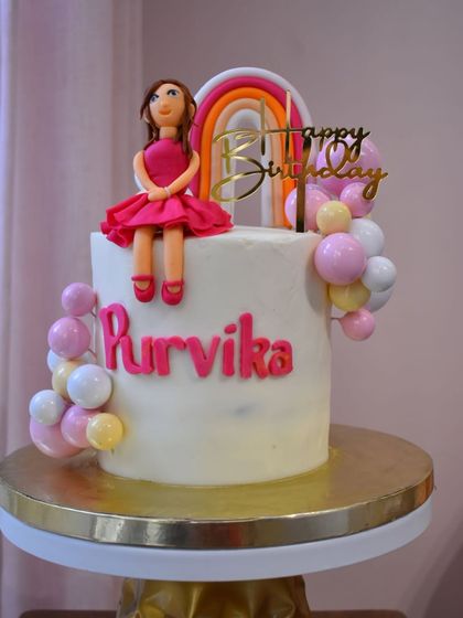 A sweet and stylish cake for Purvika's birthday. This design features a custom doll figurine in a pink dress, a rainbow topper, and a cascade of pastel-colored balloon spheres.